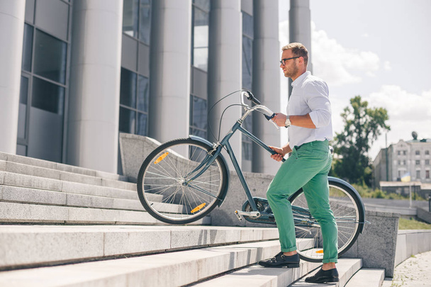 Young stylish businessman carrying bicycle up steps - Zdjęcie, obraz