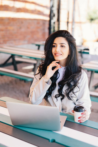 Elegant successful woman freelancer dressed formally using generic laptop computer for remote work, having rest at outdoor cafe drinking delicious coffee having dreamy expression. Lifestyle concept - 写真・画像