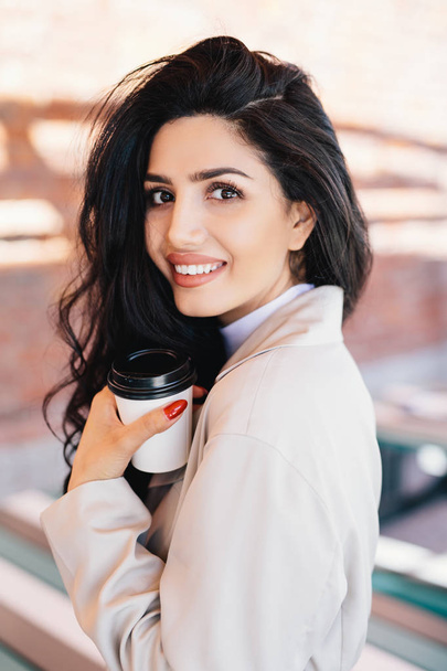 Good-looking woman with luminous dark hair, warm eyes and well-shaped lips drinking hot tea from paper cup while standing outdoors. Woman having rest enjoying drinking coffee. Rest and lifestyle - Φωτογραφία, εικόνα
