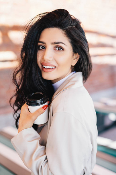 Vertical portrait of charming luxurious young lady with dark voluminous hair and appealing appearance holding takeaway coffee while waiting for her boyfriend going to have walk outdoors with him - Photo, Image