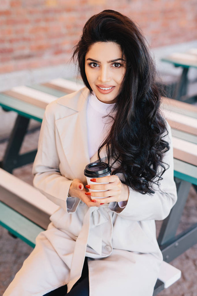 Portrait of adorable woman with dark long wavy hair, bright eyes and full lips wearing white coat sitting at bench in outdoor cafe drinking takeaway coffee posing in camera smiling pleasantly - Фото, изображение