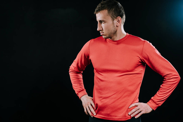 Handsome athlete man in red sportswear standing over dark background, thinking over problems in his life, holding hands on his waist, looking at one side. Lifestyle, sport, motivation. - Fotografie, Obrázek