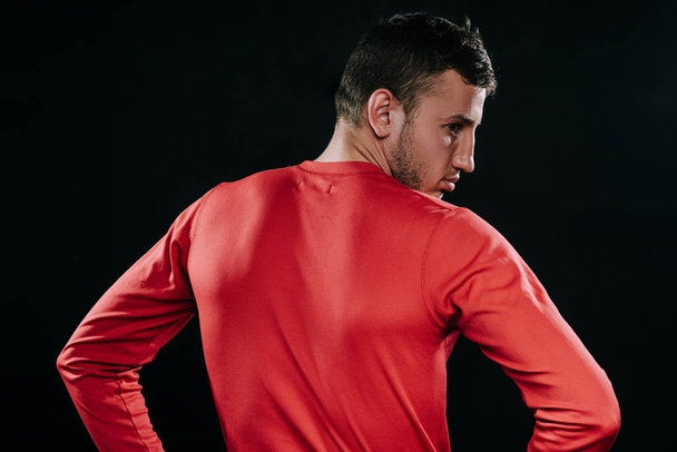 Rear view of muscular athlete man in red sportswear standing over dark background, thinking over problems in his life, holding hands on his waist, looking away. Lifestyle, sport, motivation. - Foto, Imagen