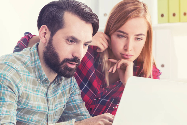 Close up of two hipsters working in a white office. A man in a green shirt is sitting at a table and pointing at a laptop screen. His blonde partner is standing near him.  - Photo, Image