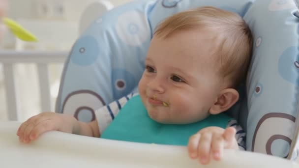 Closeup of young mother feeding cute smiling baby boy wearing bib and sitting in highchair - Felvétel, videó