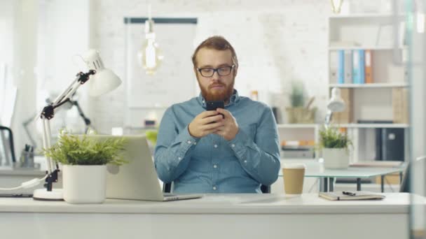 Portrait Shot of a Young Bearded Man Sitting at His Desk Using Smarphone. Open Notebook Lies on the Desk, Office is Light and Modern. - Séquence, vidéo