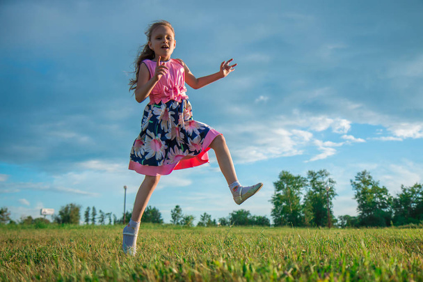Portrait of a seven-year-old girl in a bright pink dress who jumps and plays on the grass in the park on a sky background - Photo, image