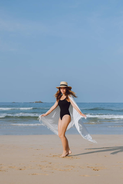Girl in a black swimsuit and hat walking and whirls on the beach  - Foto, Imagen