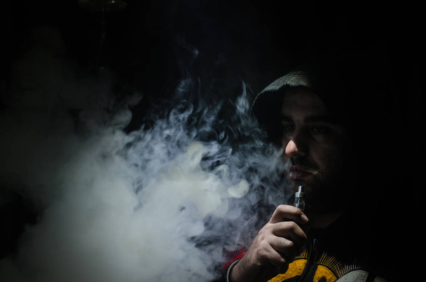 Vaping man holding a mod. A cloud of vapor. Black background. Vaping an electronic cigarette with a lot of smoke - 写真・画像
