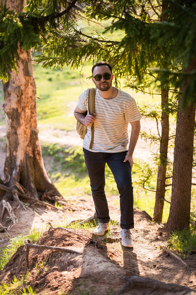 Przystojny stylowy młody człowiek sobie biały t-shirt i okulary plecak w ręku jest podróżowanie w lesie. Pozowanie na kamery w pobliżu drzewa - Zdjęcie, obraz