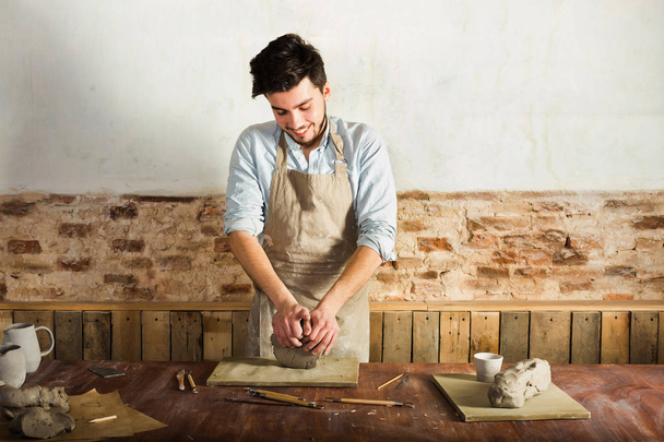 potter, workshop, ceramics art concept - standing young brunette man dressed in an apron, male hands knead the fireclay, a ceramist with raw materials on wooden table with sculpting tools set. - Foto, Imagem