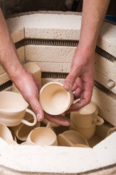 pottery, workshop, ceramics art concept - male hands gently putting the jug in the kiln, top view of electric oven for roasting of unbaked clay products, some unfinished cups and utensils. - Photo, Image