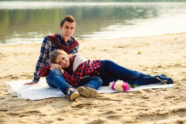 Stylish young couple in shirts and jeans while walking on the shore of a lake. A beautiful attractive girl lied on knees to her boyfriend, sitting on a plaid. - Fotoğraf, Görsel