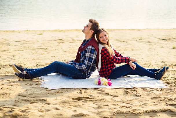 Stylish young couple in shirts and jeans while walking on the shore of a lake. Beautiful attractive girl and her handsome boyfriend leans to each other sitting on a plaid. - Foto, Imagem