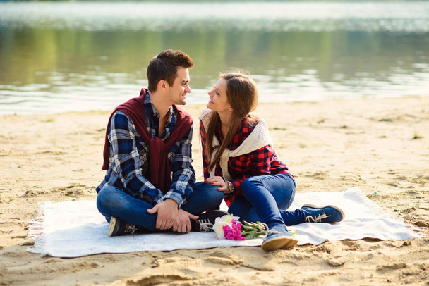 Stylish young couple in shirts and jeans while walking along the lake shore. A beautiful attractive girl and her handsome boyfriend sitting on a blanket and smiling to each other. - Foto, Imagem