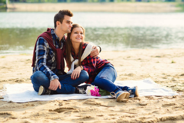 Stylish young smiling couple in shirts and jeans while walking on the shore of a lake. Beautiful attractive girl and her handsome boyfriend leans to each other sitting on a plaid. - Valokuva, kuva