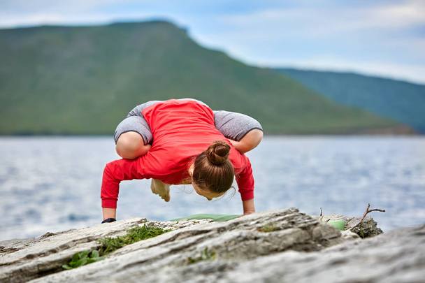 Young woman doing yoga exercises on stone near big river against beautiful landscape. - Photo, Image