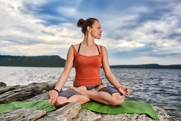 Красивая молодая женщина медитирует в позе йоги на камне возле реки
. - Фото, изображение