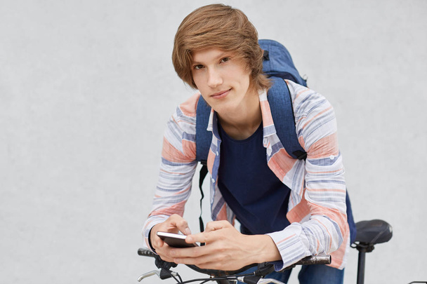 Modne uczniak trzyma plecak koszulkę za pomocą telefonów komórkowych surfowanie portali społecznościowych komunikuje się z jego przyjaciółmi, opierając się na rowerze. Stylowe hipster facet z roweru i smartphone - Zdjęcie, obraz