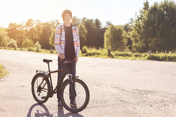 Healthy lifestyle concept. Teenage male with trendy hairstyle wearing casual shirt, jeans and sport shoes standing with bicycle on road having rest outdoors holding his bicycle by the handlebar - Фото, изображение