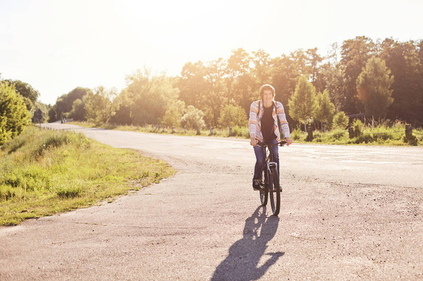 People, sports and healthy active lifestyle concept. Smiling hipster schoolboy with trendy hairdo riding bicycle outdoors. Young cyclist having vacations travelling with bicycle admiring nature - Fotoğraf, Görsel