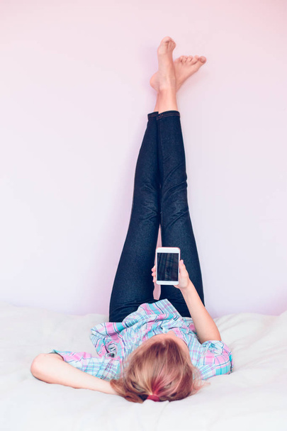 Young gril using mobile phone lying in bed with legs up - Foto, immagini