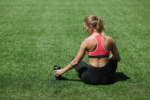 Young unrecognizable sporty woman athlete in sportswear sitting, keep water or protein cocktail shake on stadium field green grass, workout concept - Fotografie, Obrázek