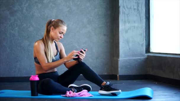 Mujer en el gimnasio con smartphone. Ella sentada en la alfombra y usando un teléfono inteligente
 - Imágenes, Vídeo