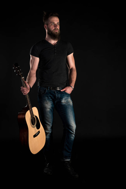 Een charismatische en stijlvolle man met een baard staat full-length met een akoestische gitaar op een geïsoleerde zwarte achtergrond. Verticale frame - Foto, afbeelding