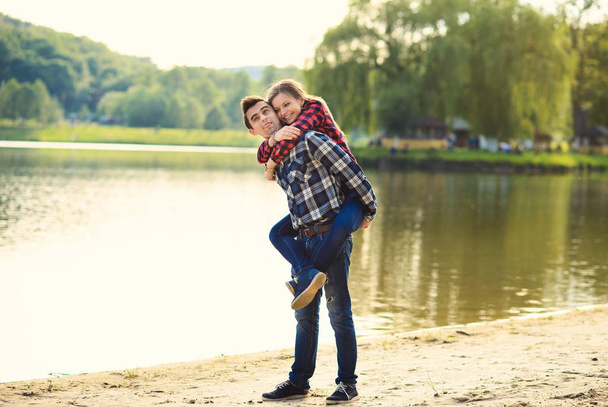 Stylish young smiling couple in shirts and jeans while walking along the lake shore. Beautiful charming girl jumped on back to her handsome boyfriend. - Foto, imagen