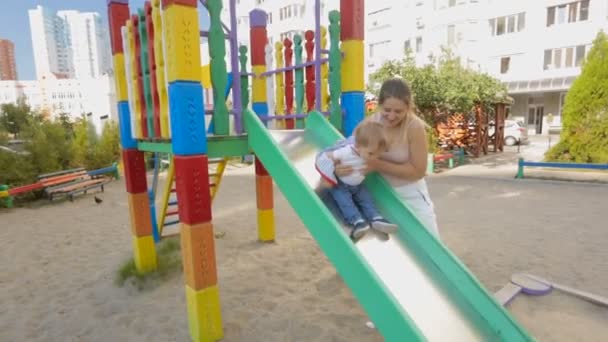 Glückliche junge Mutter hilft ihrem kleinen Sohn auf Spielplatz auf der Rutsche - Filmmaterial, Video