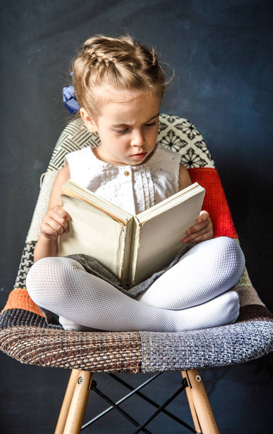 kleines Mädchen sitzt auf einem Stuhl und liest ein Buch - Foto, Bild