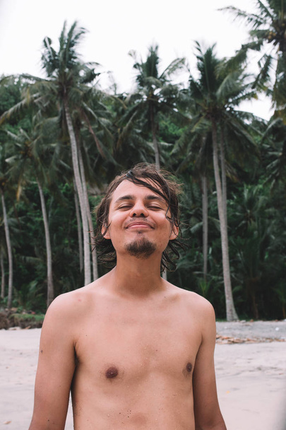 Young tanned man on the beach smiles, palm trees on the background, Indonesia, Bali - 写真・画像