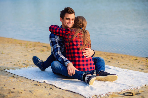 Stylish young couple in shirts and jeans while walking on the shore of a lake. Beautiful charming girl hugs her handsome boyfriend sitting on a plaid. - Foto, Imagem
