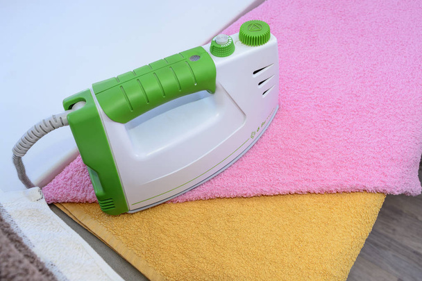 Bügelwäsche mit Bügeleisen. ein Stapel gebügelter Handtücher, die neben dem Bügeleisen liegen. Heißes Bügeleisen auf einem Bügelbrett. Teflonsohle mit kleinen Löchern bedeckt - Foto, Bild