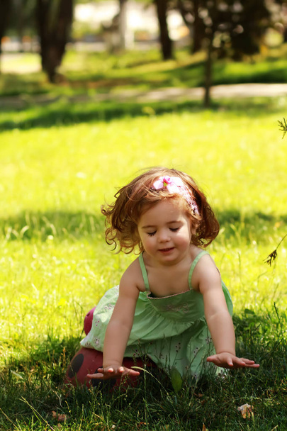 夏の光緑のドレスでかわいい 1 歳女の子が芝生の草に触れる喜びを得る  - 写真・画像