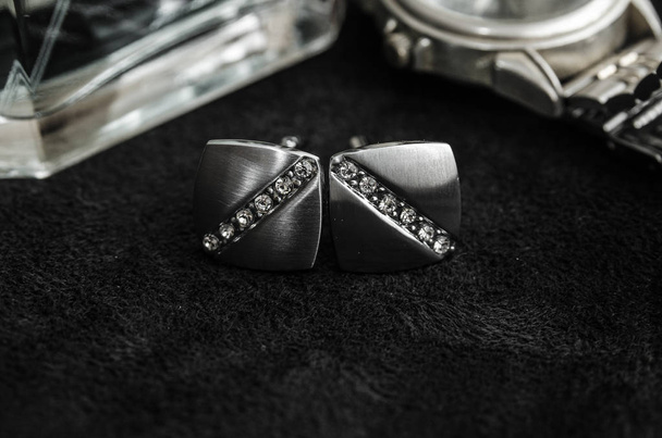 Silver cufflinks with precious stones on black matter between perfumes and watch, black and white photo - Photo, image