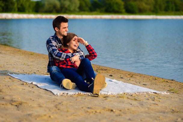 Stylish young smiling couple in shirts and jeans while walking on the shore of a lake. Handsome boyfriend hugs his beautiful charming girl  sitting on a plaid. - Fotoğraf, Görsel