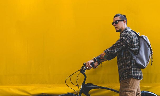 Bonito adulto homem em óculos de sol anda com uma bicicleta ao longo de uma parede amarela espaço de cópia
 - Foto, Imagem