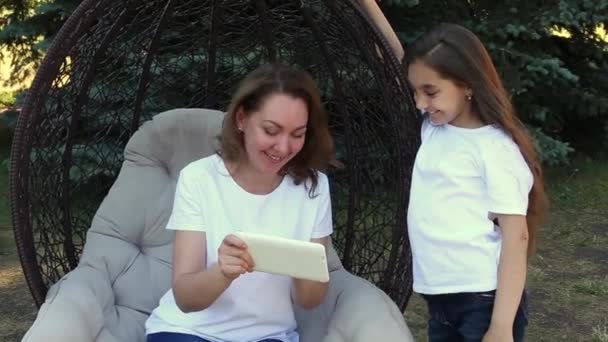 Happy mother and daughter using white tablet PC, sitting in a park at summer day - Imágenes, Vídeo