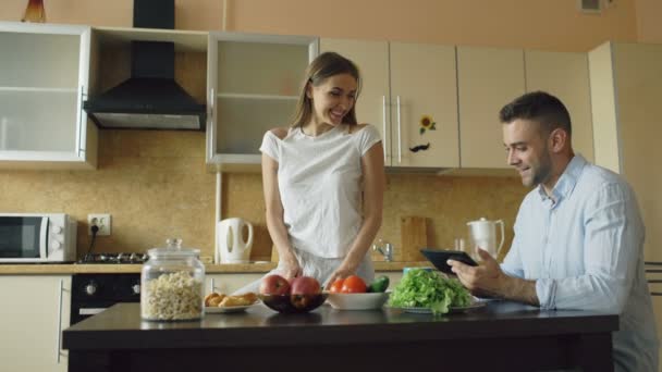 Attractive couple chatting in the kitchen early morning. Handsome man using tablet while his girlfriend cooking - Felvétel, videó