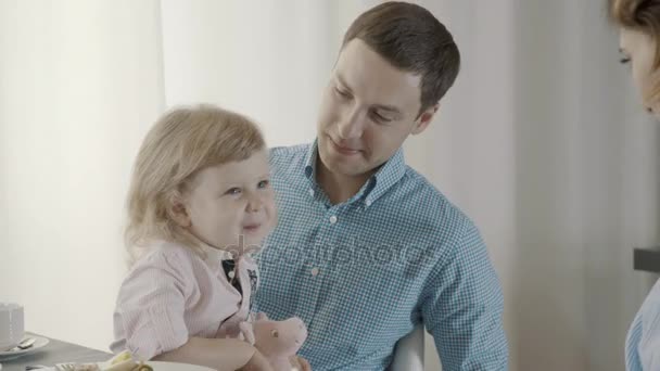 A little girl is sitting on her fathers lap, Mom is playing with her - Felvétel, videó