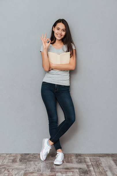 Smiling cute young woman holding book and showing ok gesture - Фото, зображення
