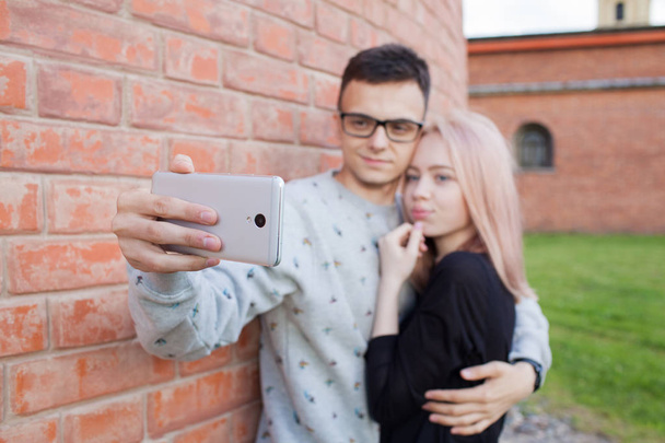 Genç bir çift bir selfie kırmızı tuğla duvar arka plan üzerinde smartphone ile fotoğraf çekmek. Mavi gözleri ve Arap görünüm kahverengi gözlü genç bir adam sarışın kız. - Fotoğraf, Görsel