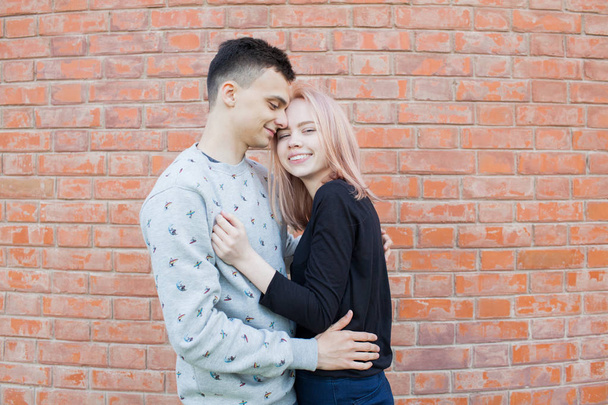 Szczęśliwa Młoda para przytulanie i uśmiecha się na tle ściany z czerwonej cegły. Blondynka, niebieskie oczy i młody człowiek Emiraty wyglądu z brązowe oczy - Zdjęcie, obraz