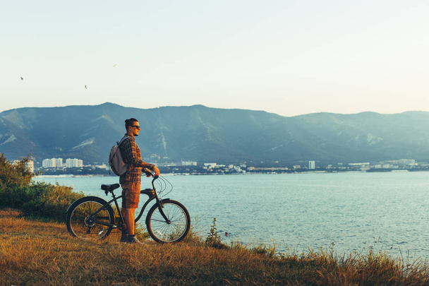 Junger Mann steht mit Fahrrad an der Küste und genießt Aussicht auf Sonnenuntergang - Foto, Bild
