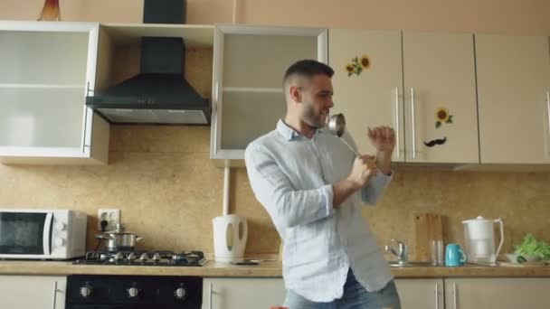 Atraktivní mladý legrační muž tančí a zpívá s naběračku při vaření v kuchyni doma - Záběry, video