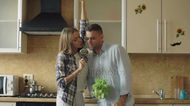 Attractive young joyful couple have fun dancing and singing while cooking in the kitchen at home - Felvétel, videó