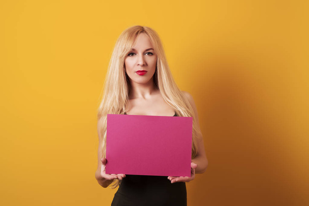 Beautiful young blond woman is holding blank board sign isolated on yellow background. - Zdjęcie, obraz