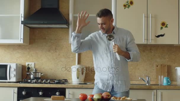 Slow motion of Attractive young funny man dancing and singing with ladle while cooking in the kitchen at home - Materiał filmowy, wideo
